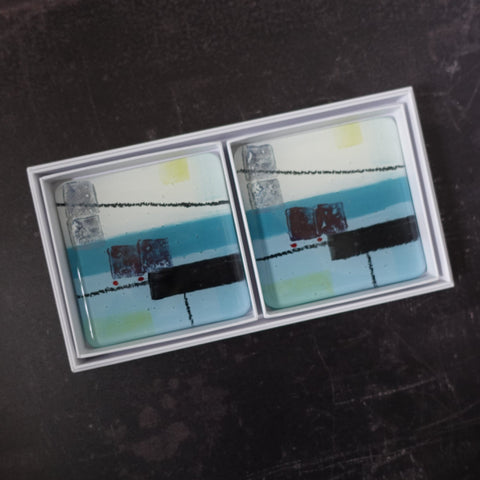Set of Two coasters  by Jenny Hoole. Set of two coasters. In ocean blues with a hint of soft yellow and abstract dark shapes in rectangles suggesting a spring tide in a harbour. Fused glass drinks mat in a luxury white gift box