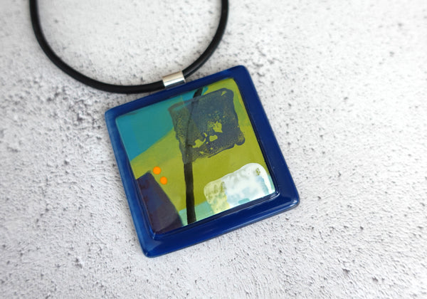 5cm square glass pendant on a 45cm rubber necklace. Navy blue base and green, cyan and white pattern hand painted in enamel
