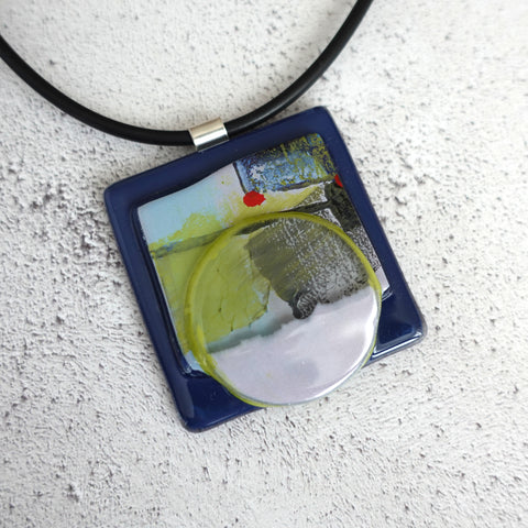 A square 5cm glass pendant. Hand painted with enamels in an abstract pattern. Red, lime green and navy. Sterling silver bail and rubber necklace