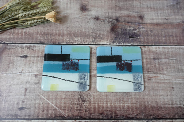 Abstract Spring Tide coasters by Jenny Hoole. Blocks of deep blue colours over a light blue and cream background with a hint of soft yellow . 10 x 10cm with bumper feet underneath